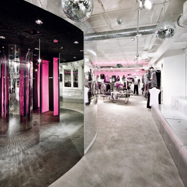 A modern clothing store with a large mirrored wall, featuring a disco ball and colorful clothes displayed on racks and tables.