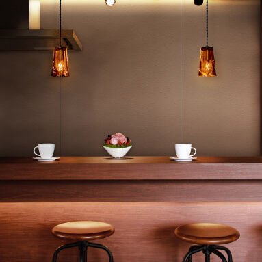 Dark brown wooden bar with two cups of coffee and a fruit bowl. Two amber pendant lights hang above the bar. The wall behind the bar is light brown.