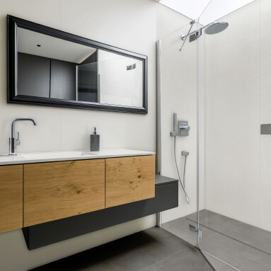 Baño moderno con mueble bajo lavabo suspendido de madera natural, un gran espejo con marco negro y una cabina de ducha de vidrio. El panel de pared con acabado de madera aporta calidez y comodidad al espacio.