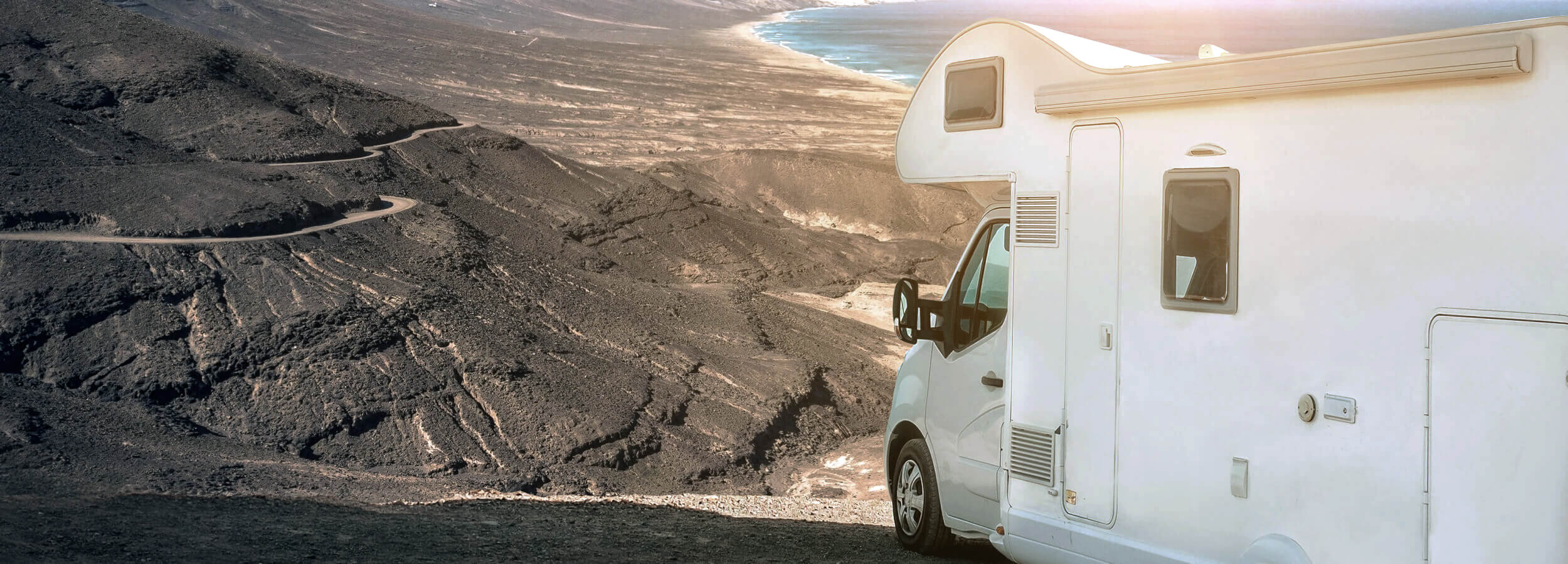 Una autocaravana blanca aparcada en un acantilado con vistas a una carretera sinuosa que conduce a un vasto océano azul.