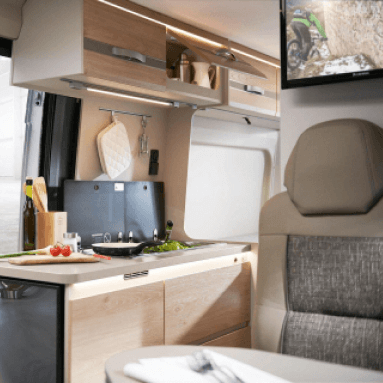 Cómoda zona de cocina en la autocaravana con un panel de pared de madera. El panel se utiliza para fijar estantes, ganchos y otros elementos, creando espacio de almacenamiento adicional para utensilios de cocina. La encimera está hecha de un material claro, lo que amplía visualmente el espacio. En la encimera hay una estufa, un fregadero y otros electrodomésticos necesarios para cocinar.