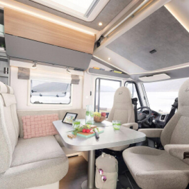 Una sala de estar espaciosa y luminosa en la autocaravana, decorada con un estilo moderno. Una acogedora zona de estar con una mesa plegable, una gran ventana y un techo panorámico crean un ambiente cómodo y permiten disfrutar del viaje.