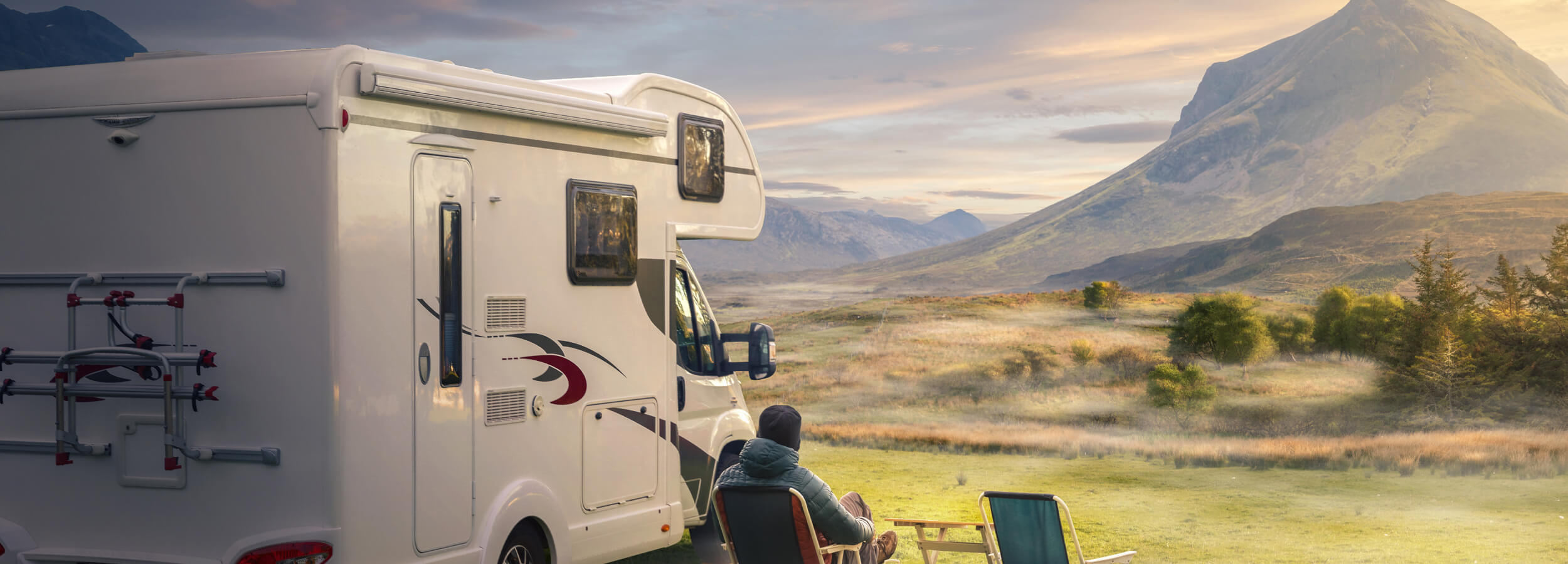 Un hombre está sentado en una silla plegable frente a una autocaravana blanca estacionada en un campo. Al fondo, hay una cordillera y el cielo está pintado con los cálidos colores del atardecer.