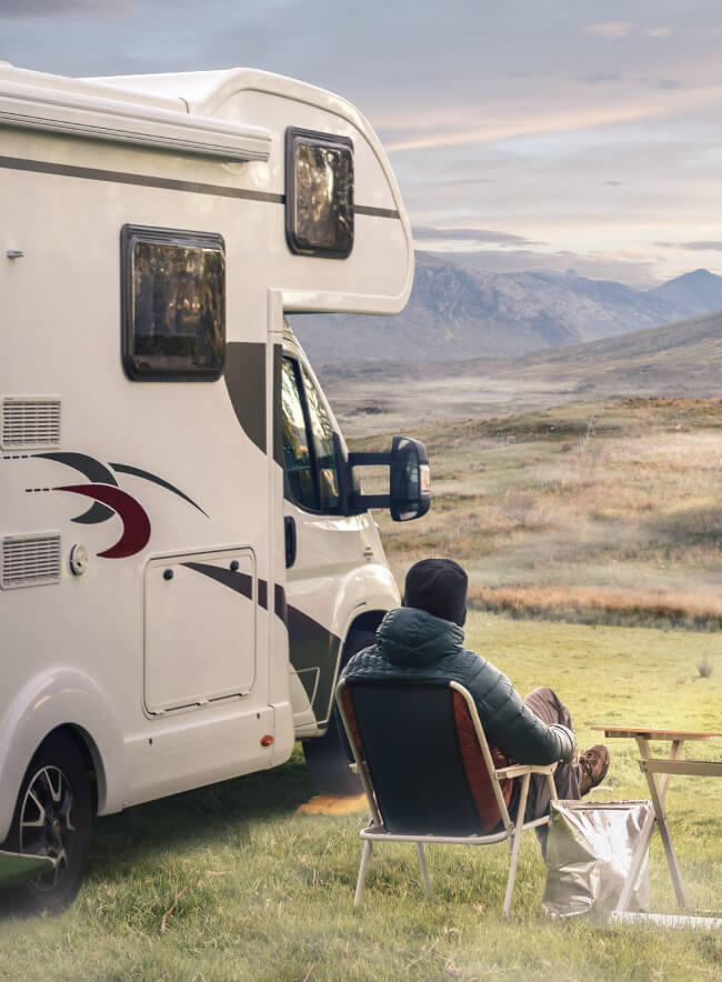 Un hombre está sentado en una silla plegable frente a una autocaravana blanca estacionada en un campo. Al fondo, hay una cordillera y el cielo está pintado con los cálidos colores del atardecer.