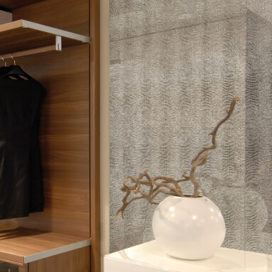 Elegante interior de un vestidor: un panel de pared con una textura de cuero natural crea un acento de lujo sobre el fondo de armarios de madera. Un jarrón blanco con ramas añade un toque de naturaleza.