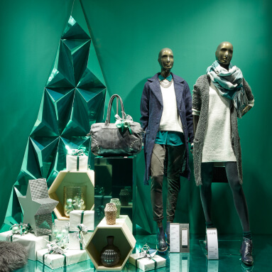 Esta imagen presenta un escaparate festivo con dos maniquíes vestidos con moda de invierno, de pie frente a un panel de pared con acabado metálico. El diseño geométrico reflectante del panel de pared imita la forma de un árbol de Navidad, agregando un toque moderno y lujoso a la escena. Los maniquíes están vestidos con capas acogedoras, como abrigos, bufandas y botas, perfectas para una colección de invierno. Alrededor de ellos, cajas de regalo, adornos y objetos decorativos están dispuestos para realzar el tema festivo. El panel de pared metálico agrega profundidad y elegancia a la exhibición, creando una visual llamativa para la ventana de una tienda de moda.