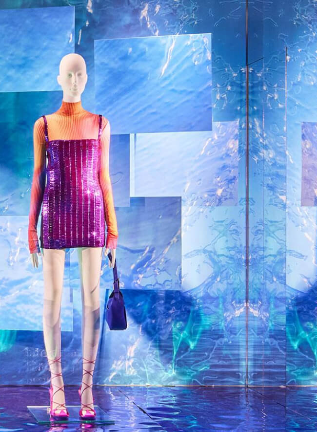 Un escaparate de una tienda de moda muestra un maniquí con un vestido vibrante frente a un panel de pared abstracto. La superficie espejada del panel, con su efecto metálico, crea una ilusión de profundidad y refleja la luz ambiental, amplificando el impacto visual. La combinación inusual de colores y texturas llama la atención y subraya el diseño moderno de la colección.