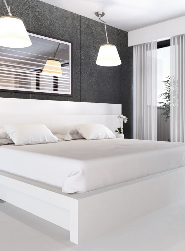 Dormitorio moderno con una pared de piedra oscura, una cama blanca con sábanas y almohadas blancas crujientes, dos lámparas colgantes, un cuadro enmarcado y un balcón con cortinas blancas y vistas a los árboles. La habitación está llena de luz natural.