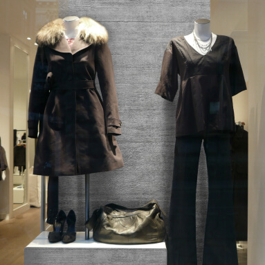 Vitrina de una tienda de moda con ropa de mujer. En primer plano, maniquíes presentan un abrigo marrón con cuello de piel y un conjunto negro compuesto por una blusa y pantalones. El fondo de la vitrina está diseñado en un estilo contemporáneo con un panel de hormigón, creando un acento industrial.