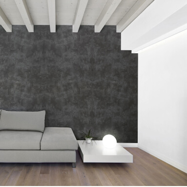 Un salón moderno con una pared de acento acabada para parecer concreto. El panel oscuro y texturizado contrasta con el suelo de madera clara y las vigas del techo blancas, creando un ambiente elegante y acogedor. El sofá gris y la mesa de café blanca completan el diseño minimalista.