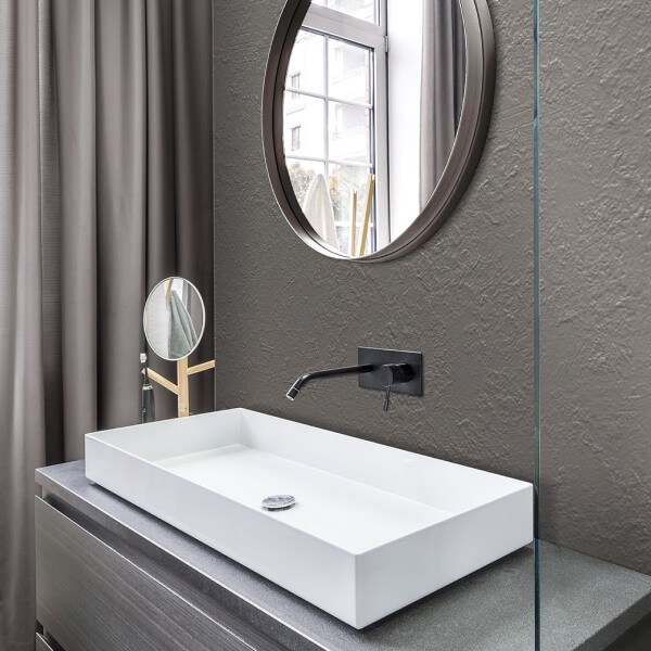 Un baño moderno con un elegante panel de pared gris que crea un fondo texturizado. Un lavabo de cerámica blanca sobre una encimera de madera oscura y un espejo redondo en un marco negro complementan el diseño minimalista.