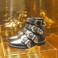 Un par de botas de cuero negro con hebillas doradas, elegantemente expuestas en un escaparate sobre un fondo de mosaico de espejo.