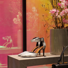 Un escaparate con un par de tacones altos plateados con una correa negra, exhibidos en una mesa. El escaparate está decorado con un panel metálico con efecto espejo, que refleja la luz rosa y naranja del exterior.