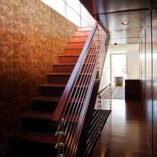 Una escalera moderna con escalones de madera y baranda de metal. Detrás de la escalera hay un panel de pared con una superficie metálica texturizada.
