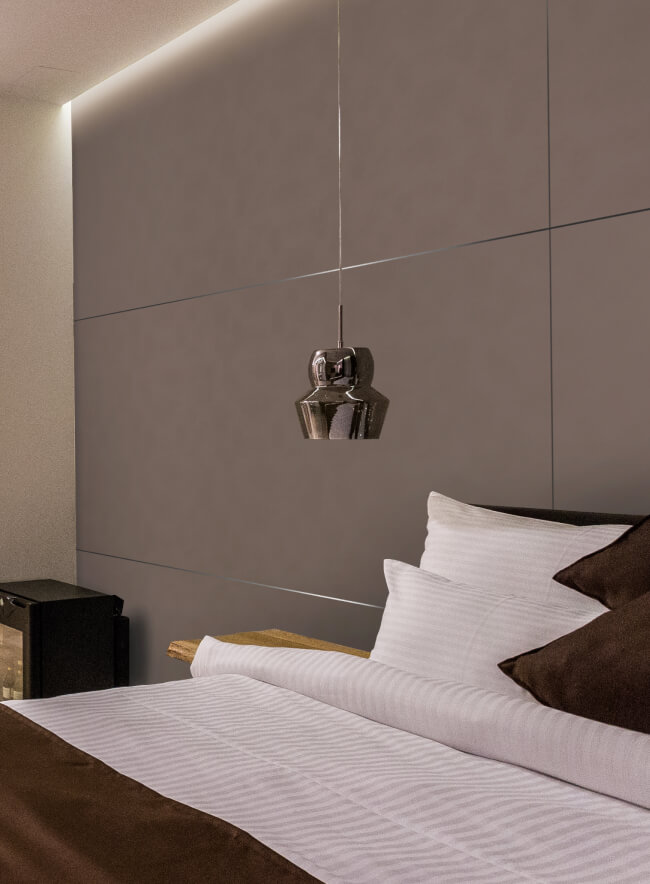 Una habitación de hotel moderna con una cama king-size, una cabina de ducha de vidrio y una pared de acento de ladrillo. La cama está vestida con sábanas blancas crujientes y almohadas marrones. La habitación está iluminada por una cálida luz superior y una lámpara de noche. La pared detrás de la cama está cubierta por un elegante panel de pared de imitación de cuero.
