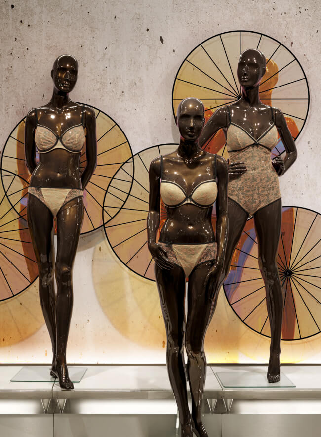 Un escaparate con tres maniquíes que llevan ropa interior elegante, frente a un panel de pared de piedra. La ropa interior se presenta en varios estilos y colores. La atmósfera general de la imagen es cálida y acogedora.
