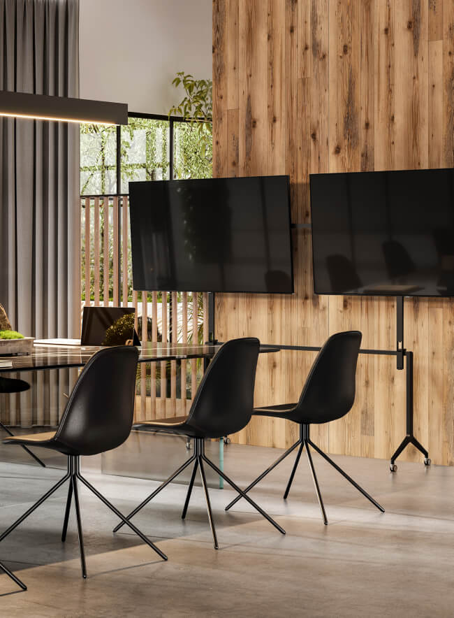 Una sala de conferencias moderna con un panel de pared de madera, una mesa de vidrio y sillas negras. Dos grandes pantallas están montadas en la pared.