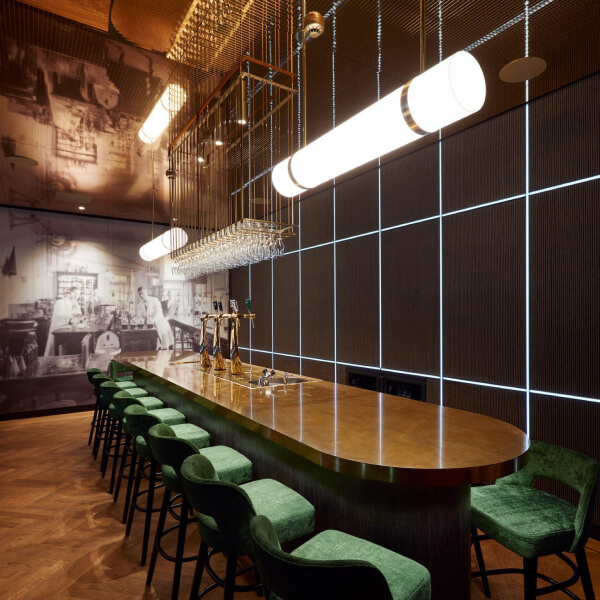 El interior de un bar elegante con una larga barra de madera oscura y sillas de terciopelo verde. En la pared detrás de la barra hay un gran cuadro que representa un proceso histórico de elaboración de cerveza. Las paredes están decoradas con pequeños mosaicos, creando una sensación de profundidad y textura. Las lámparas colgantes con pantallas mate iluminan la barra, creando un ambiente acogedor.