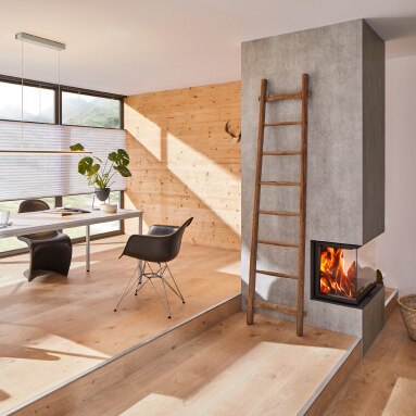 Salón moderno con chimenea, suelo de madera y pared de hormigón. La chimenea está empotrada en la pared y tiene un frontal de vidrio para que se puedan ver las llamas. Junto a la chimenea hay una escalera que conduce a un nivel superior de la habitación. La habitación está amueblada con muebles modernos, incluyendo una mesa, sillas y una planta.