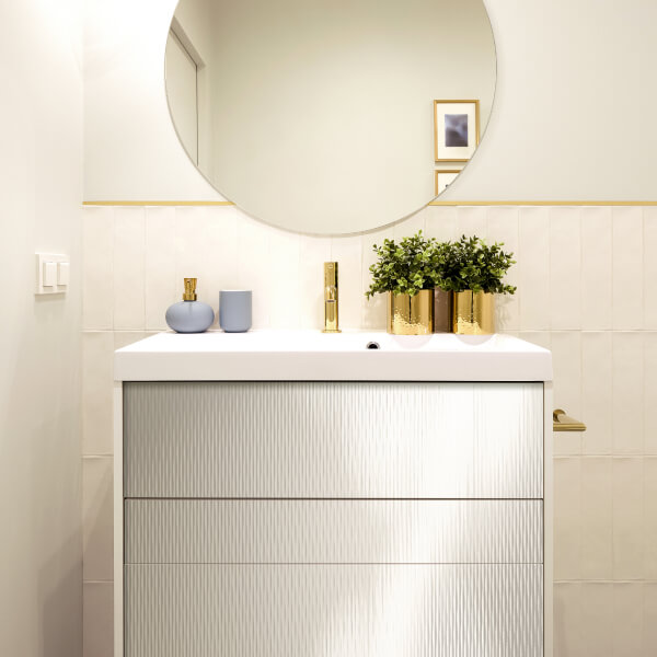 Baño moderno con una pared de acento revestida con un panel de pared de plástico texturizado. La superficie estriada del panel crea un efecto visual interesante y armoniza con los muebles y sanitarios blancos. Un espejo redondo en un marco dorado y accesorios dorados añaden lujo y elegancia al interior.