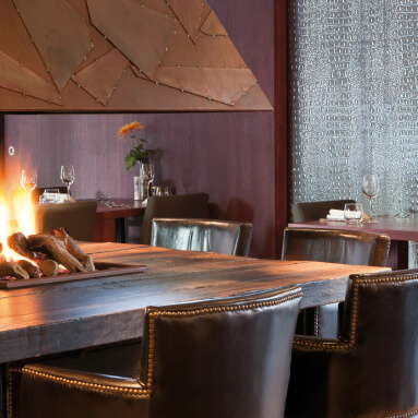 Un restaurante acogedor con una gran mesa de madera rodeada de sillas de cuero. En el centro de la mesa hay una chimenea con fuego. La pared detrás de la chimenea está decorada con un panel texturizado de imitación de piel de reptil.