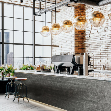 El interior de una cafetería moderna con una amplia barra acabada en piedra natural. En la pared trasera se instala un elegante panel de pared con imitación de madera, creando un ambiente acogedor. Grandes ventanales panorámicos llenan la habitación de luz natural, mientras que las lámparas colgantes con elementos metálicos añaden un encanto industrial. En la barra, hay una máquina de café, una vitrina con pasteles y altos taburetes de bar. En general, la imagen transmite la atmósfera de un establecimiento de restauración moderno y a la moda.