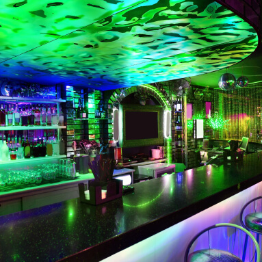 Un bar con una pared espejada con un patrón de camuflaje metálico y 3D en verde y azul neón. El bar está iluminado con luces de colores y la barra está llena de botellas de alcohol.