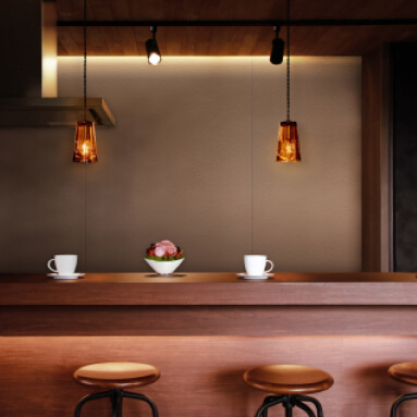 Barra de madera marrón oscuro con dos tazas de café y un bol de fruta. Dos lámparas colgantes de color ámbar cuelgan sobre la barra. La pared detrás de la barra es de color marrón claro.