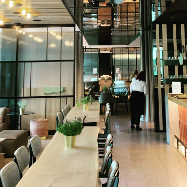 Esta imagen muestra un espacio de comedor moderno en un restaurante o cafetería con un diseño limpio y minimalista. Una larga mesa de madera con plantas verdes en macetas es la pieza central, rodeada de sillas suaves y acolchadas. A la izquierda, hay cómodas áreas de asientos, mientras que a la derecha se encuentra una barra de bar con iluminación suave. El espacio se define por sus elegantes paneles de pared con espejos, que añaden profundidad y elegancia al interior gastronómico. Los paneles de espejo reflejan la distribución abierta y aireada de la sala, mejorando su estética contemporánea. En general, el ambiente es cálido, elegante y acogedor, perfecto para comer en compañía.