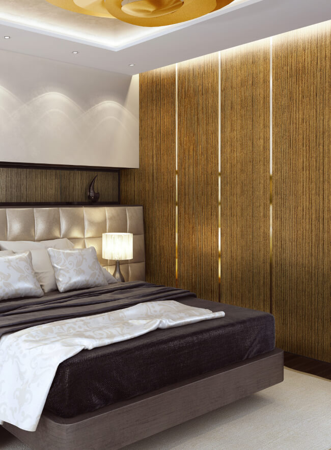 Un dormitorio moderno con un panel de pared de madera detrás de la cama, una cabecera de cuero beige y dos lámparas de noche. La cama está vestida con un edredón gris oscuro y almohadas blancas. Un jarrón de flores se encuentra en la mesita de noche.