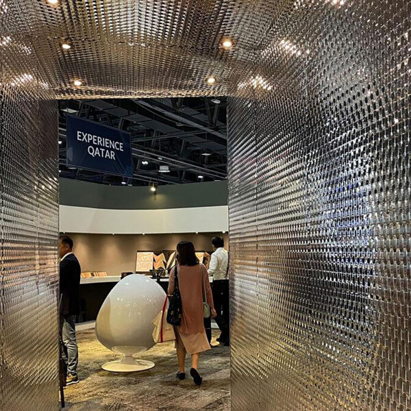 Visitantes en un stand de exposición que muestra un diseño moderno con un panel de pared estructural elegante y reflectante que parece de metal. La imagen presenta una exhibición para "Experience Qatar", con un panel de pared futurista y espejado que crea una entrada llamativa.