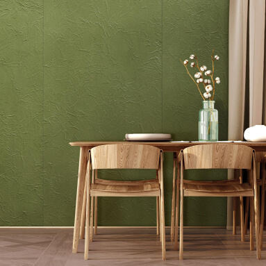 Un panel de pared texturizado de cuero sintético de un verde intenso. El panel está instalado en un comedor, donde hay una larga mesa de madera con cuatro sillas de madera y un jarrón con ramas de algodón. El suelo es de madera oscura.