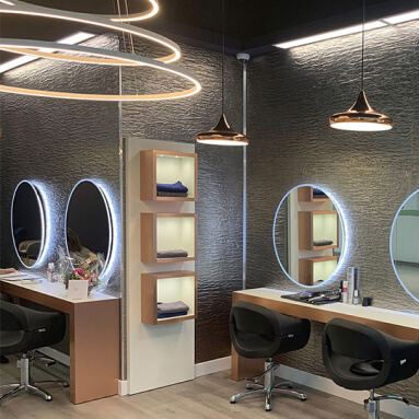 Una peluquería moderna con un panel de pared de metal texturizado, tres espejos redondos y un estante de madera con toallas.