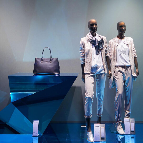 Esta imagen muestra un escaparate de moda con dos maniquíes vestidos con atuendos modernos de temática metálica. Están posicionados frente a un elegante panel de pared espejado que agrega un toque moderno y lujoso a la presentación. Los maniquíes están estilizados con ropa ligera y reflectante, con elementos casuales pero elegantes, como bufandas y tops con patrones, transmitiendo un aspecto urbano pulido. A la izquierda, una mesa de exhibición geométrica metálica sostiene un elegante bolso de cuero, iluminado con suave luz azul. El panel de pared reflectante realza la estética futurista y de alta gama de la exhibición, convirtiéndola en un elemento visual llamativo para la ventana de una tienda de moda.