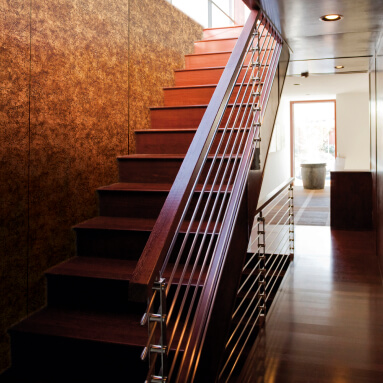 Eine moderne Treppe aus dunklem Holz mit einem Geländer aus rostfreiem Stahl führt zu einer rustikalen Metallwand mit einer Textur, die an Rost erinnert.